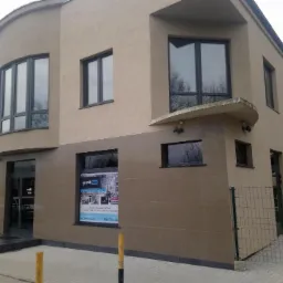 Budynek firmy kamieniarskiej z elewacją częściowo wyłożoną płytami kamiennymi w kolorze beżowym, widoczny plakat reklamowy w oknie.