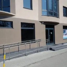Elewacja budynku z beżową okładziną, szarym cokołem i rampą dla niepełnosprawnych z metalową balustradą. Widoczne okna z ciemnymi ramami i szklane drzwi wejściowe. Na elewacji widoczna reklama...