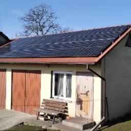 Dom z panelami fotowoltaicznymi na dachu, widok na garaż z dwoma bramami i ławką przed oknem, w tle drzewo bez liści.