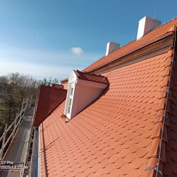 Przemysław Szuszkiewicz - Czerwony dach z dachówki karpiówki, lukarny z białymi obramowaniami, kominy i rusztowania w tle, błękitne niebo. Widok z góry na ukończony dach.