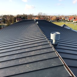 Przemysław Szuszkiewicz - Nowoczesny, grafitowy dach z blachy na budynku z wentylacją kominkową. W tle zabudowa jednorodzinna i błękitne niebo. Widok z góry.