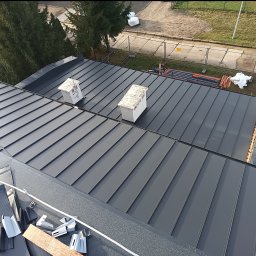 Przemysław Szuszkiewicz - Szary dach panelowy na budynku z dwoma kominami wentylacyjnymi, widok z góry. Elementy montażowe na pierwszym planie. Usługi dekarskie w Kotli.