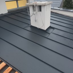 Przemysław Szuszkiewicz - Szary dach z blachy na rąbek stojący, z widocznym kominem i elementami instalacji odgromowej. Widoczne krople deszczu na powierzchni dachu.