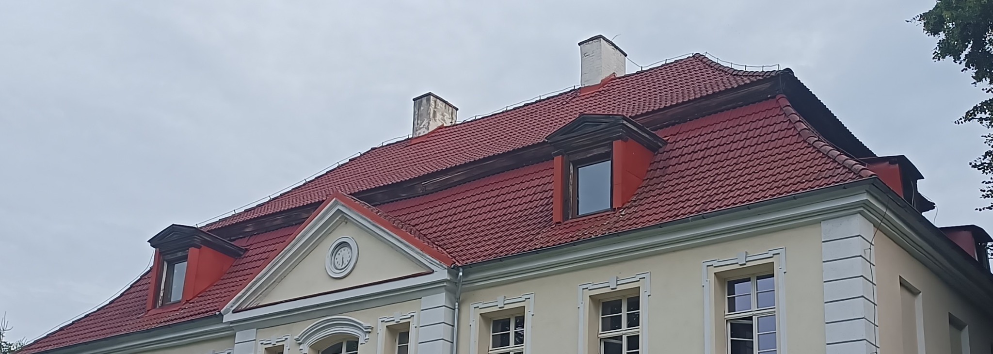 Czerwony dach z dachówką karpiówką na budynku z oknami dachowymi i dwoma kominami. Na szczycie ściany frontowej zegar.