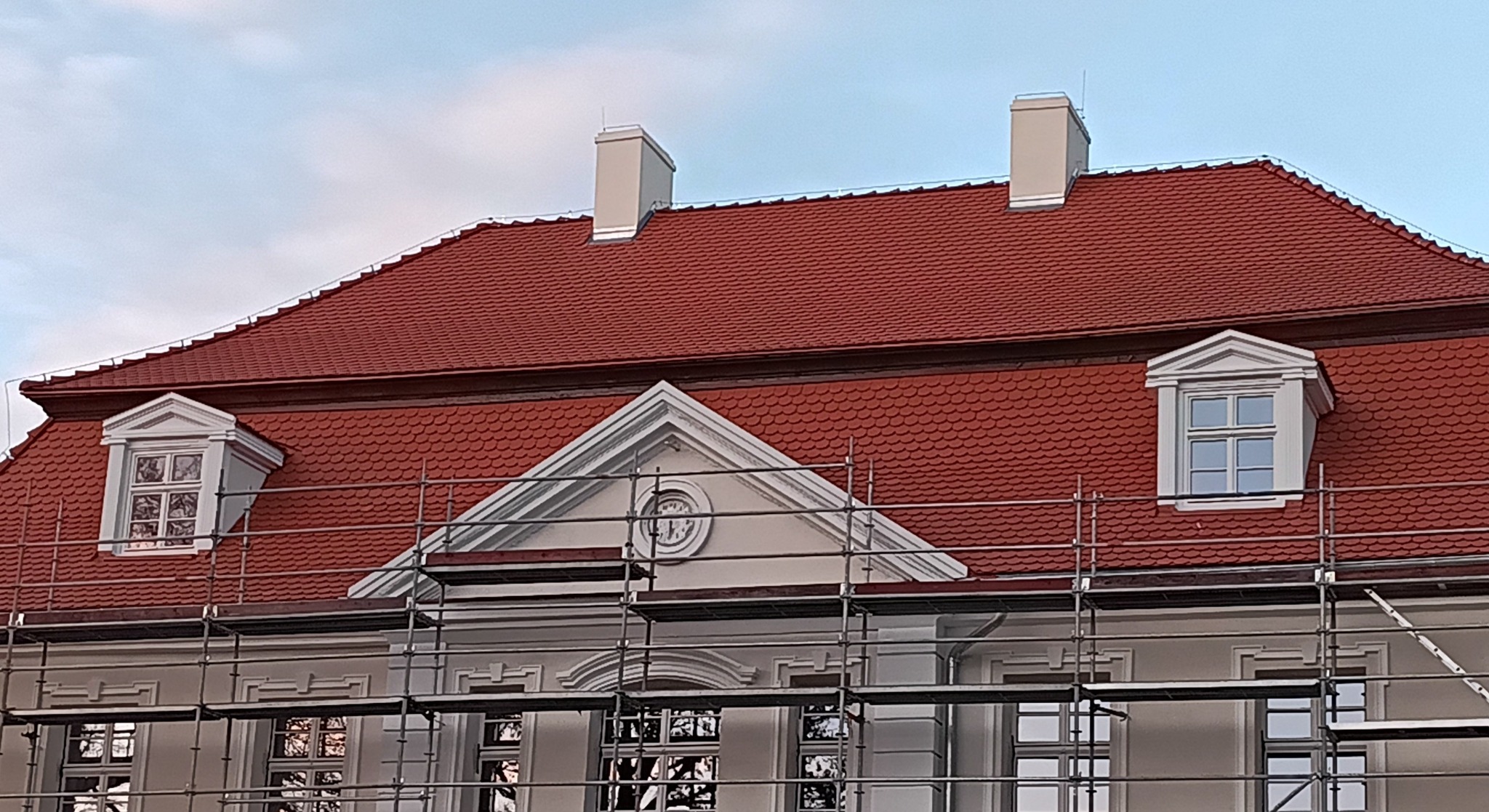 Czerwony dach z lukarnami na budynku w stylu klasycystycznym, częściowo zasłonięty rusztowaniem. Widoczne kominy i detale architektoniczne.