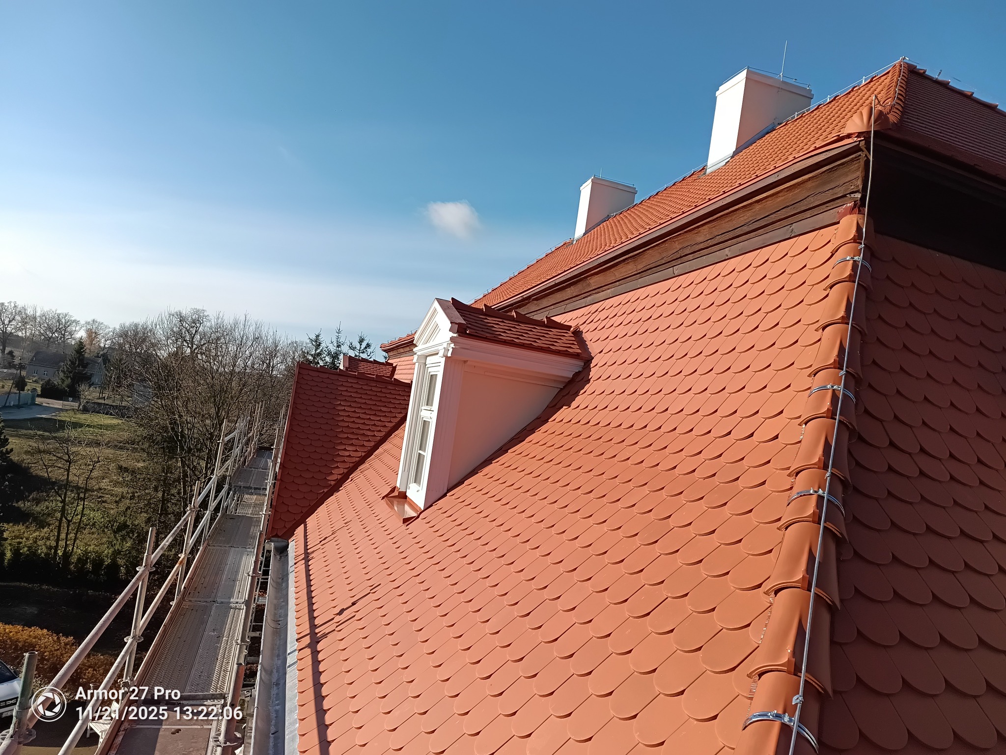 Czerwony dach z dachówki karpiówki, lukarny z białymi obramowaniami, kominy i rusztowania w tle, błękitne niebo. Widok z góry na ukończony dach.