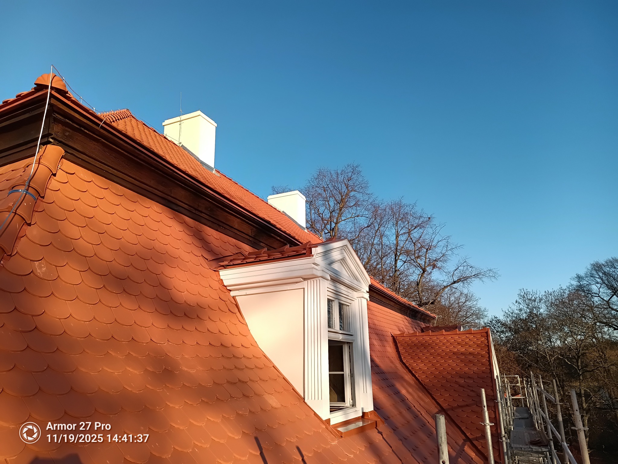 Czerwony dach z lukarną i białymi kominami na tle błękitnego nieba. Widoczne rusztowania i drzewa bez liści. Dach pokryty dachówką karpiówką.