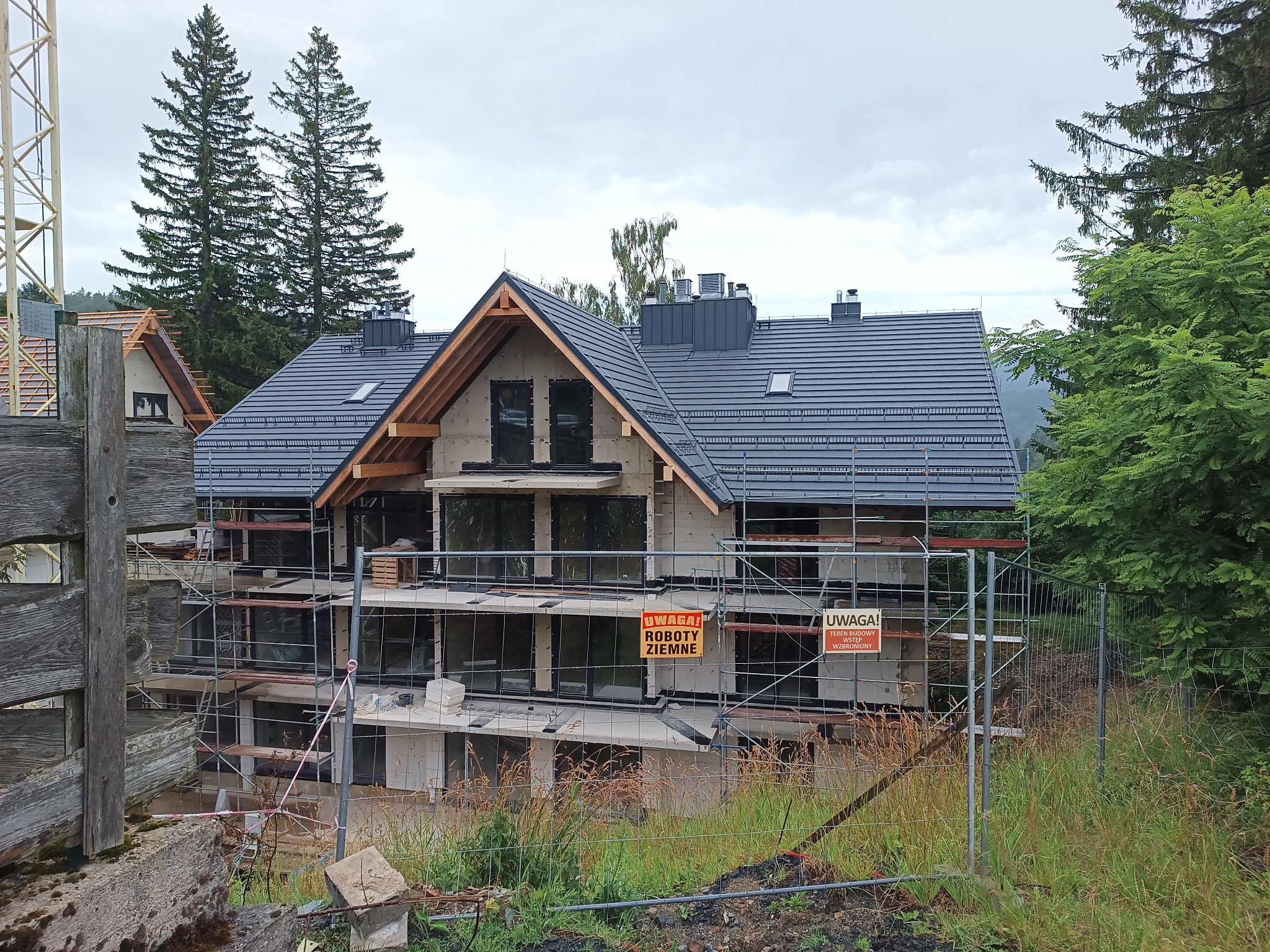 Dom w budowie z szarą dachówką, otoczony rusztowaniami i ogrodzeniem. Widoczne elementy konstrukcji dachu i kominy. W tle drzewa iglaste i liściaste.