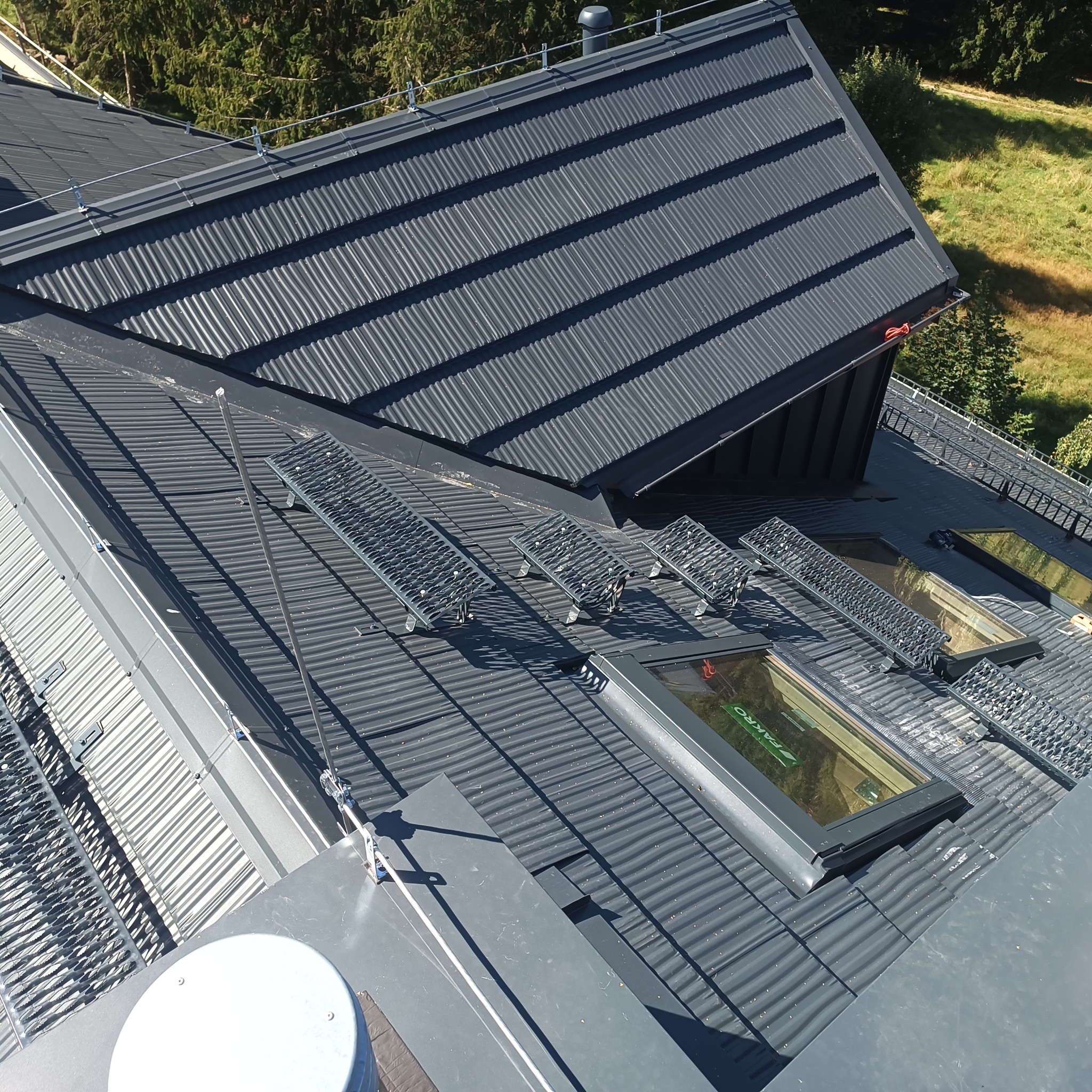 Szary dach z blachy trapezowej z oknami dachowymi i metalowymi stopniami kominiarskimi. Widok z góry, oświetlenie słoneczne.