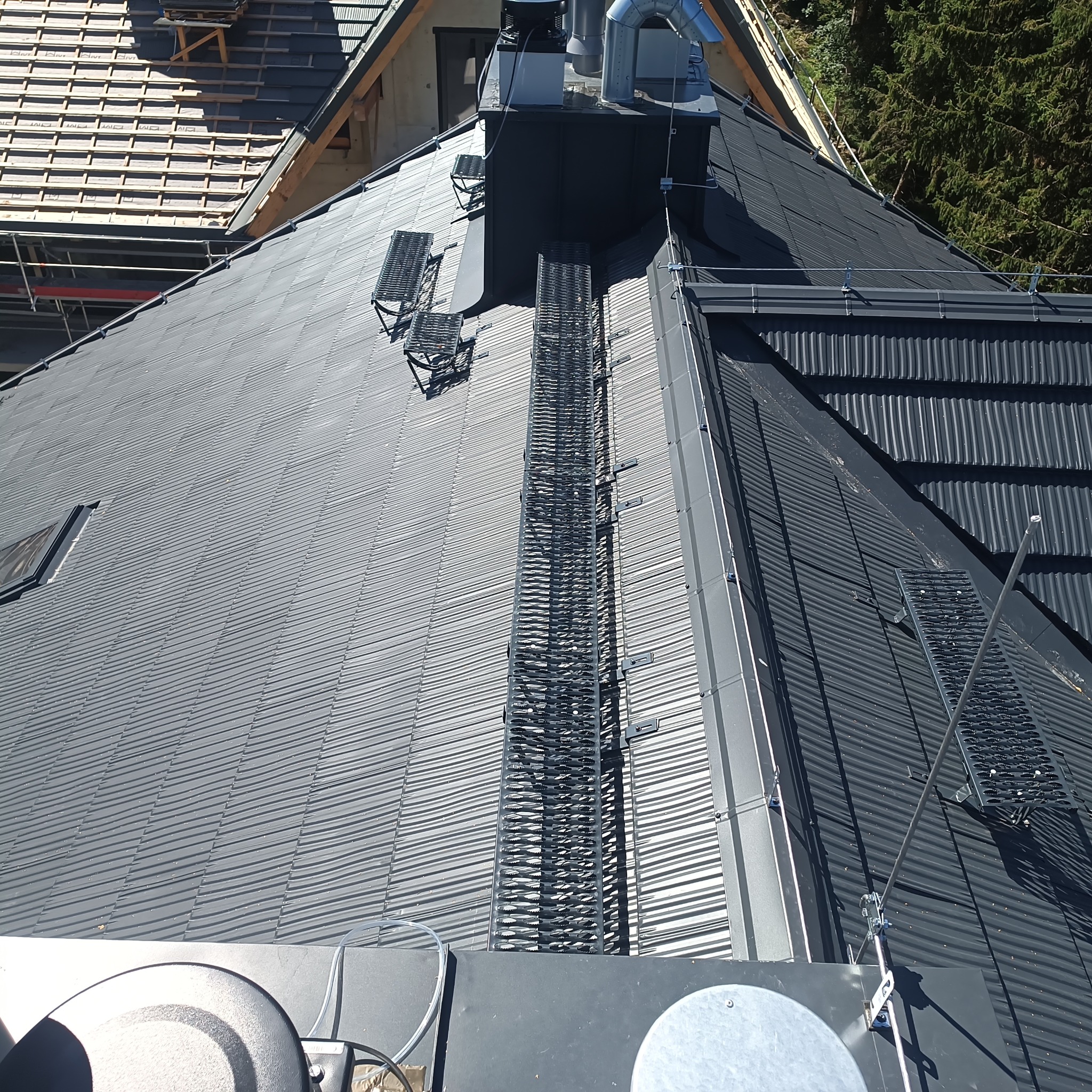 Szary, blaszany dach z zamontowanymi stopniami kominiarskimi i zabezpieczeniem przeciwśniegowym. Widok z góry na fragment dachu i komin.