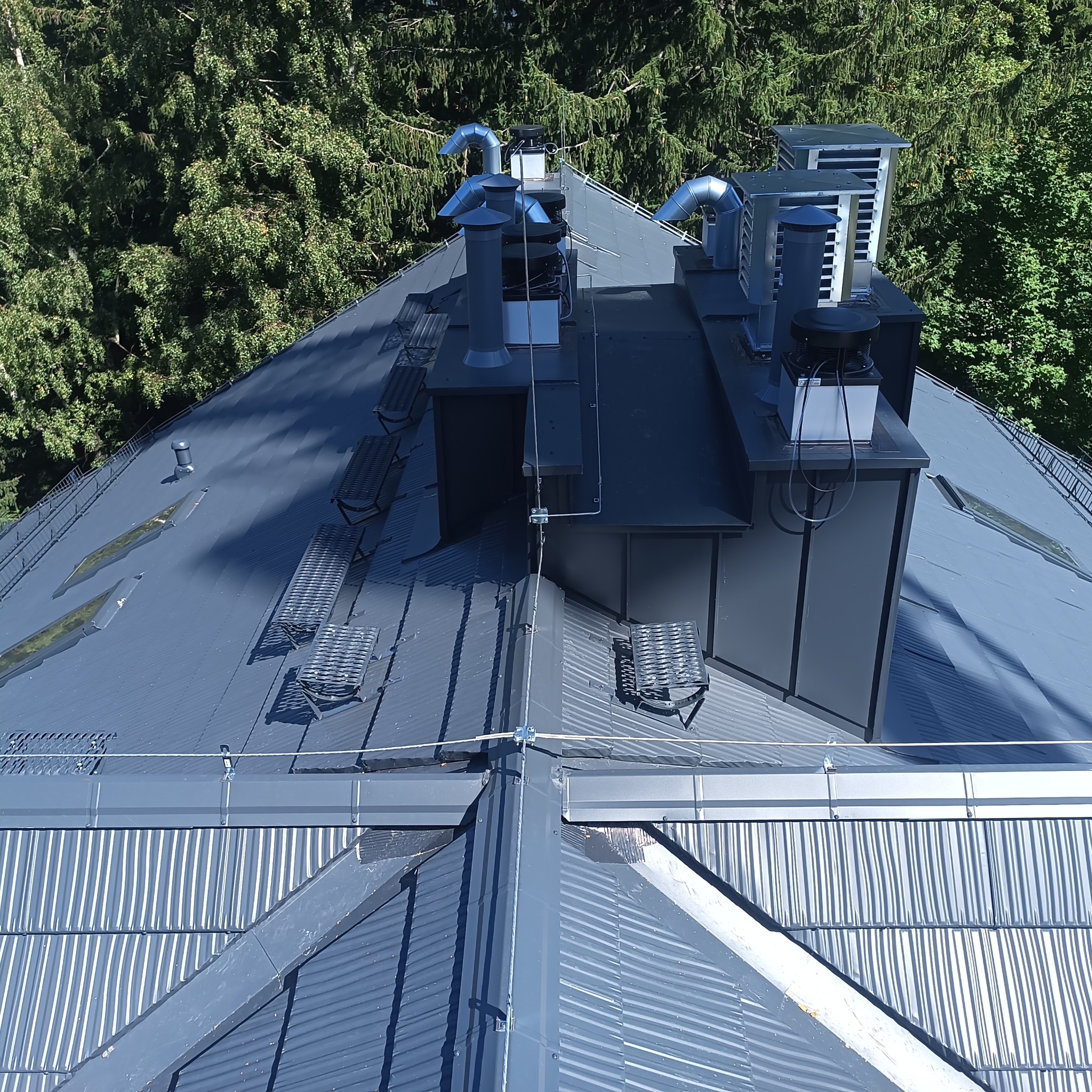 Szary dach z blachy trapezowej z kominami i zabezpieczeniami przeciwśniegowymi, widok z góry na tle drzew. Widoczne detale łączeń blachy i mocowania odgromu.