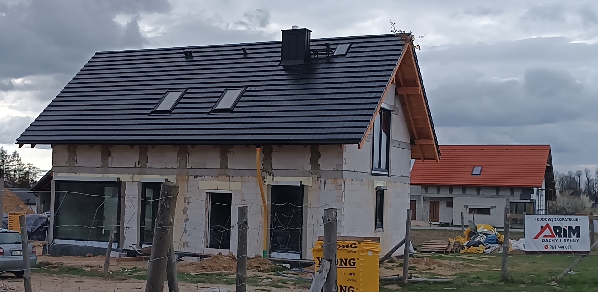 Dom w trakcie budowy z czarnym dachem z dachówki ceramicznej i oknami połaciowymi. Widoczny drugi budynek z czerwonym dachem w tle. Pochmurne niebo.