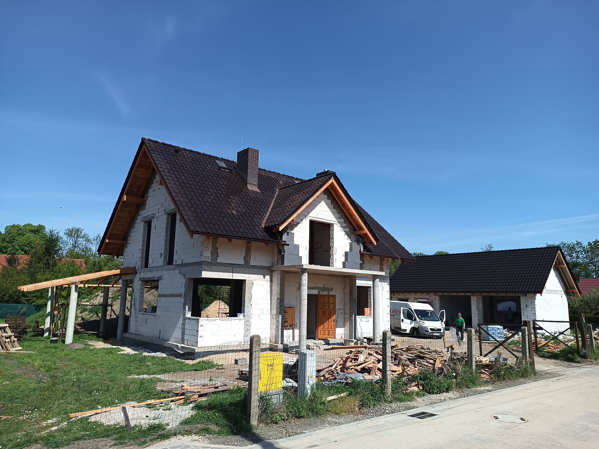 Dom w trakcie budowy z ciemnym dachem i widocznymi elementami konstrukcyjnymi. Na pierwszym planie porozrzucane drewno i częściowo widoczny płot.