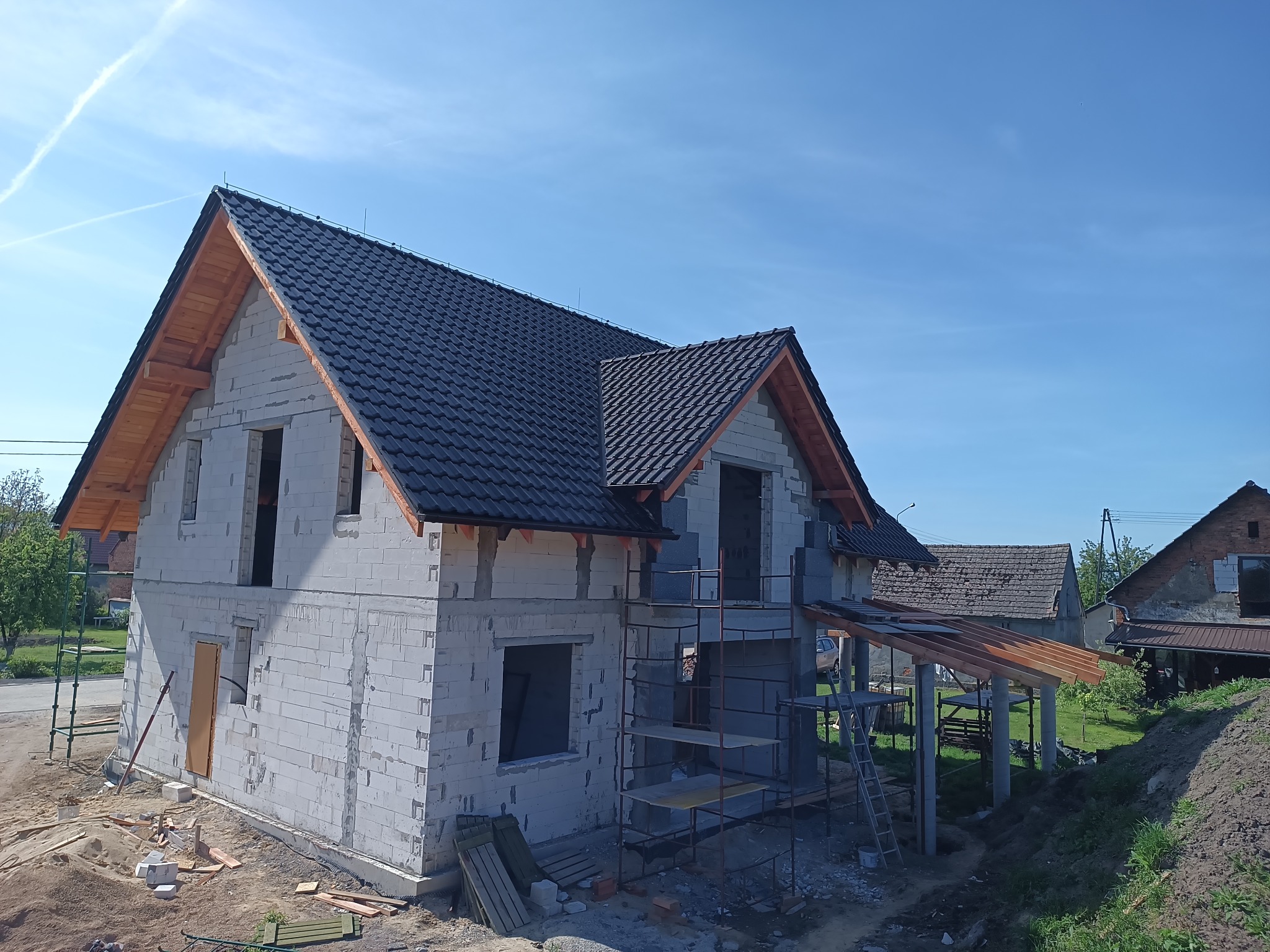 Dom w trakcie budowy z ciemną dachówką i drewnianą konstrukcją dachu. Widoczne rusztowania i materiały budowlane. Błękitne niebo w tle.