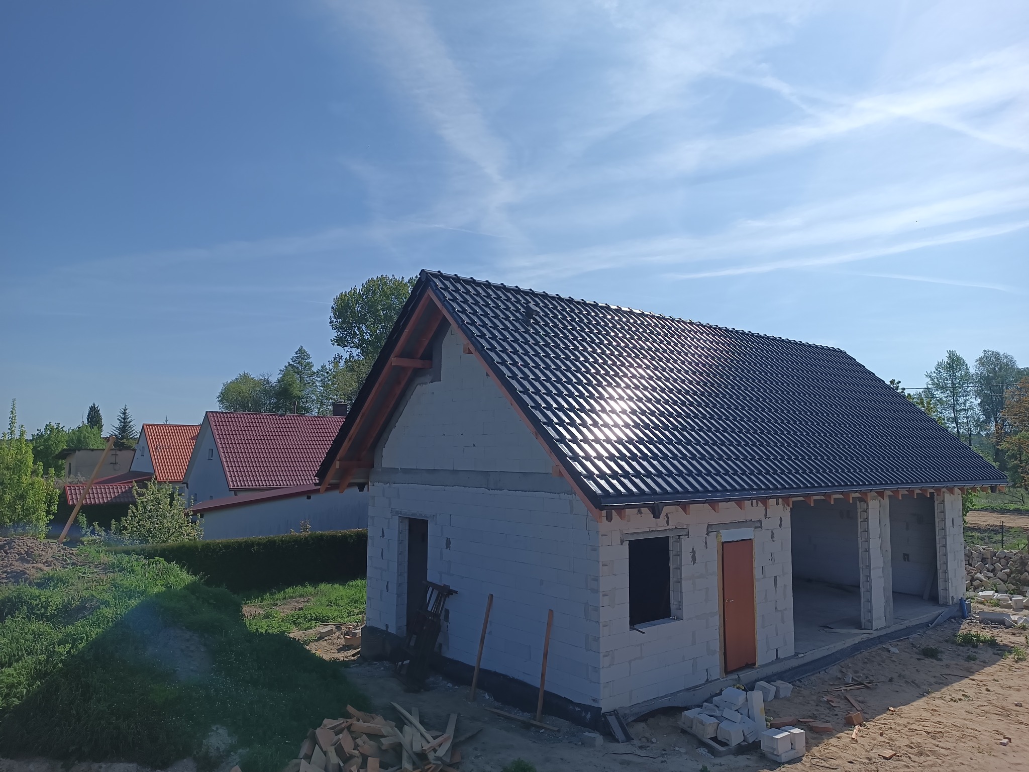 Dom w trakcie budowy z czarnym dachem w świetle słonecznym. Widoczne bloczki betonowe, drewniane elementy konstrukcji dachu i fragmenty innych budynków w tle.