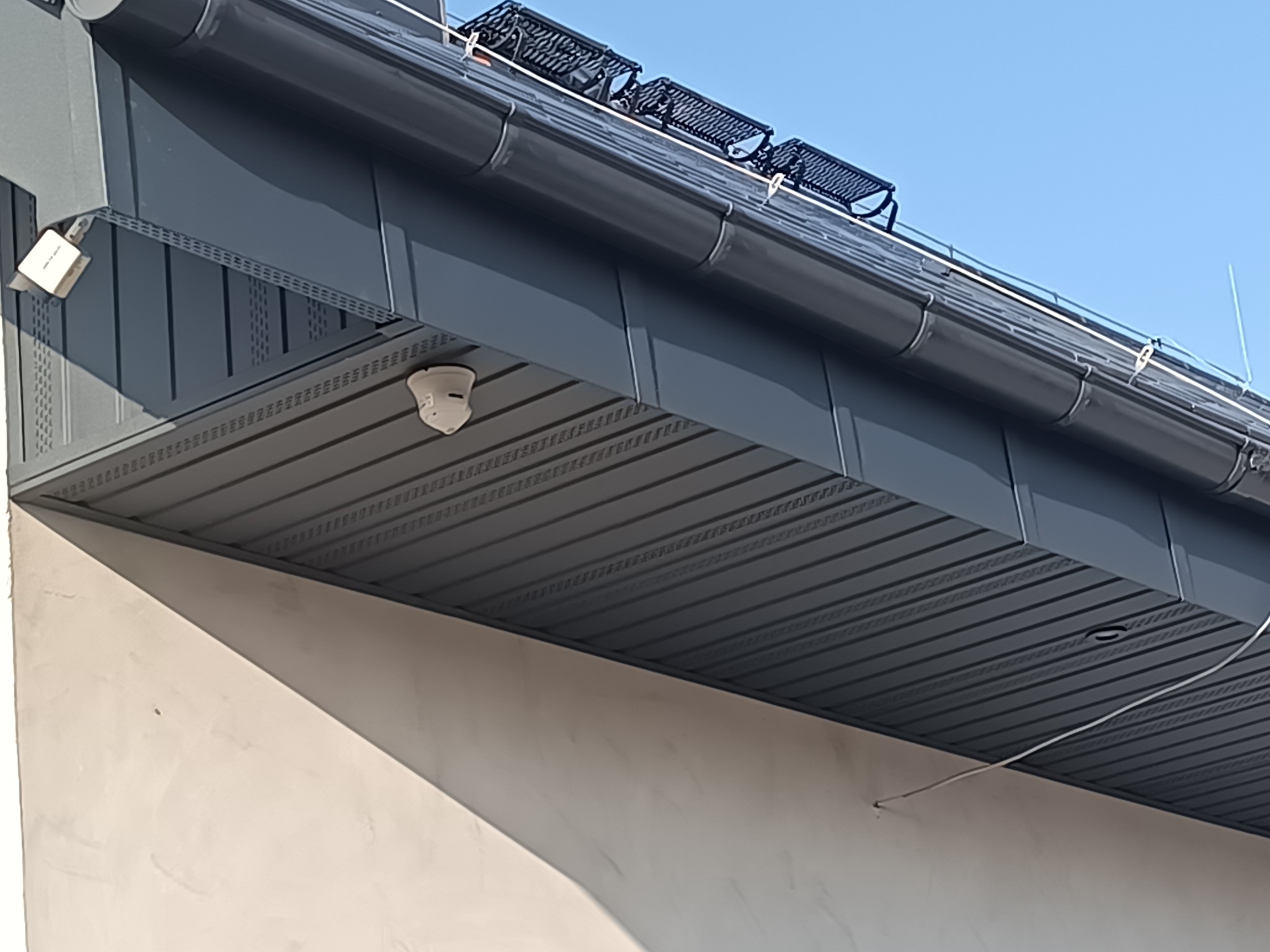 Szara podbitka dachowa z oświetleniem punktowym i rynnami w nowoczesnym domu. Widoczne elementy mocujące i fragment elewacji w jasnym odcieniu.