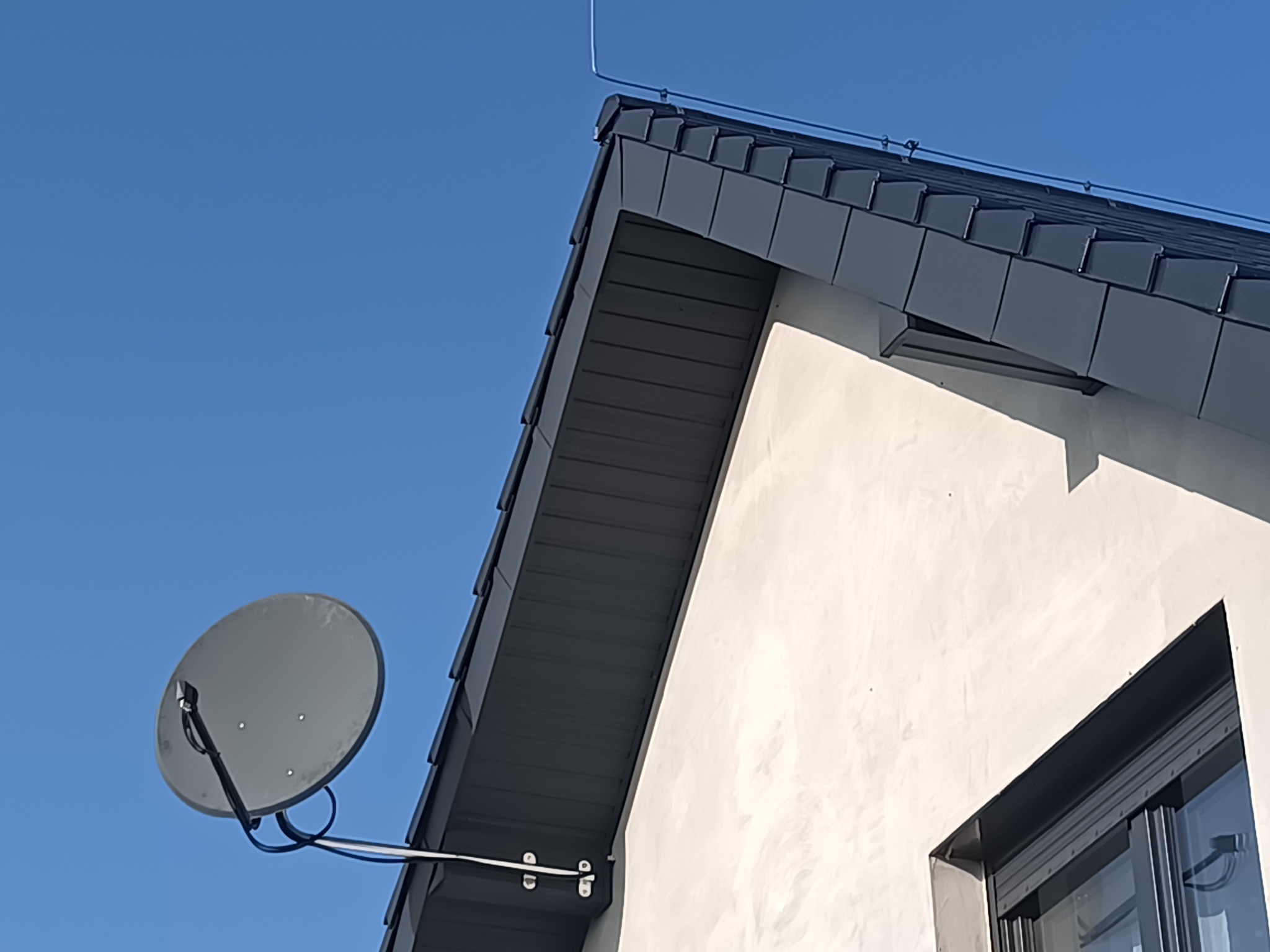 Szary dach z blachy na jasnym budynku, widoczna antena satelitarna i odgromnik. Ujęcie z dołu, błękitne niebo w tle.