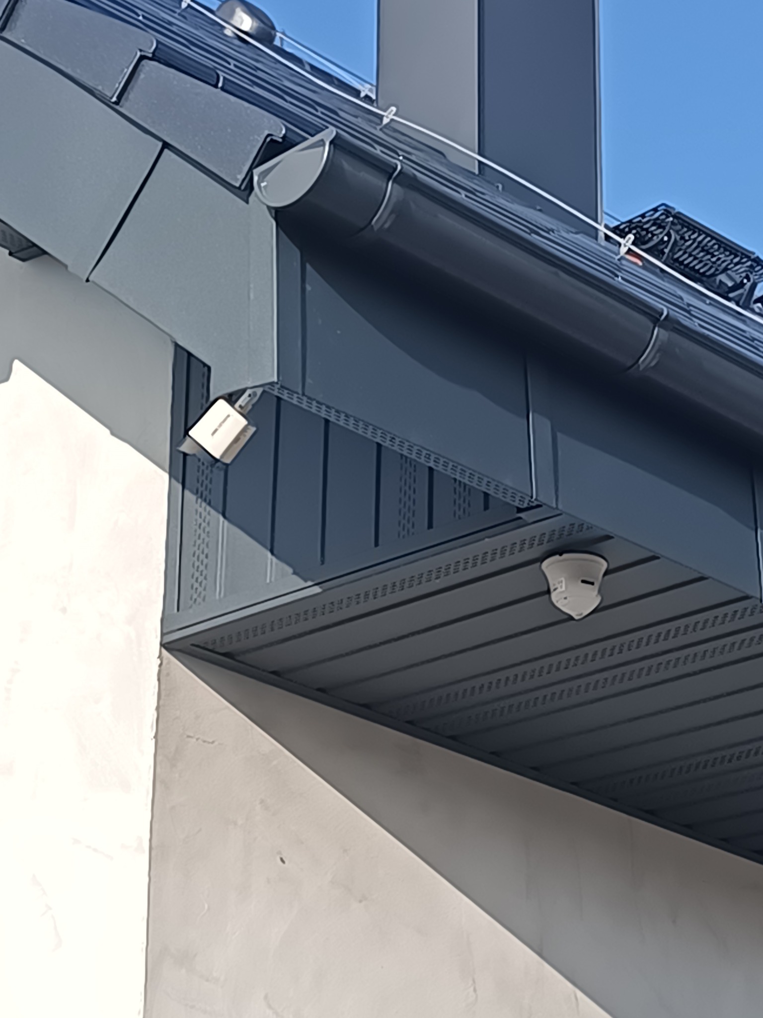 Szczegółowe ujęcie fragmentu dachu z ciemną dachówką, rynną i elementami wykończeniowymi, oświetlonym naturalnym światłem słonecznym.