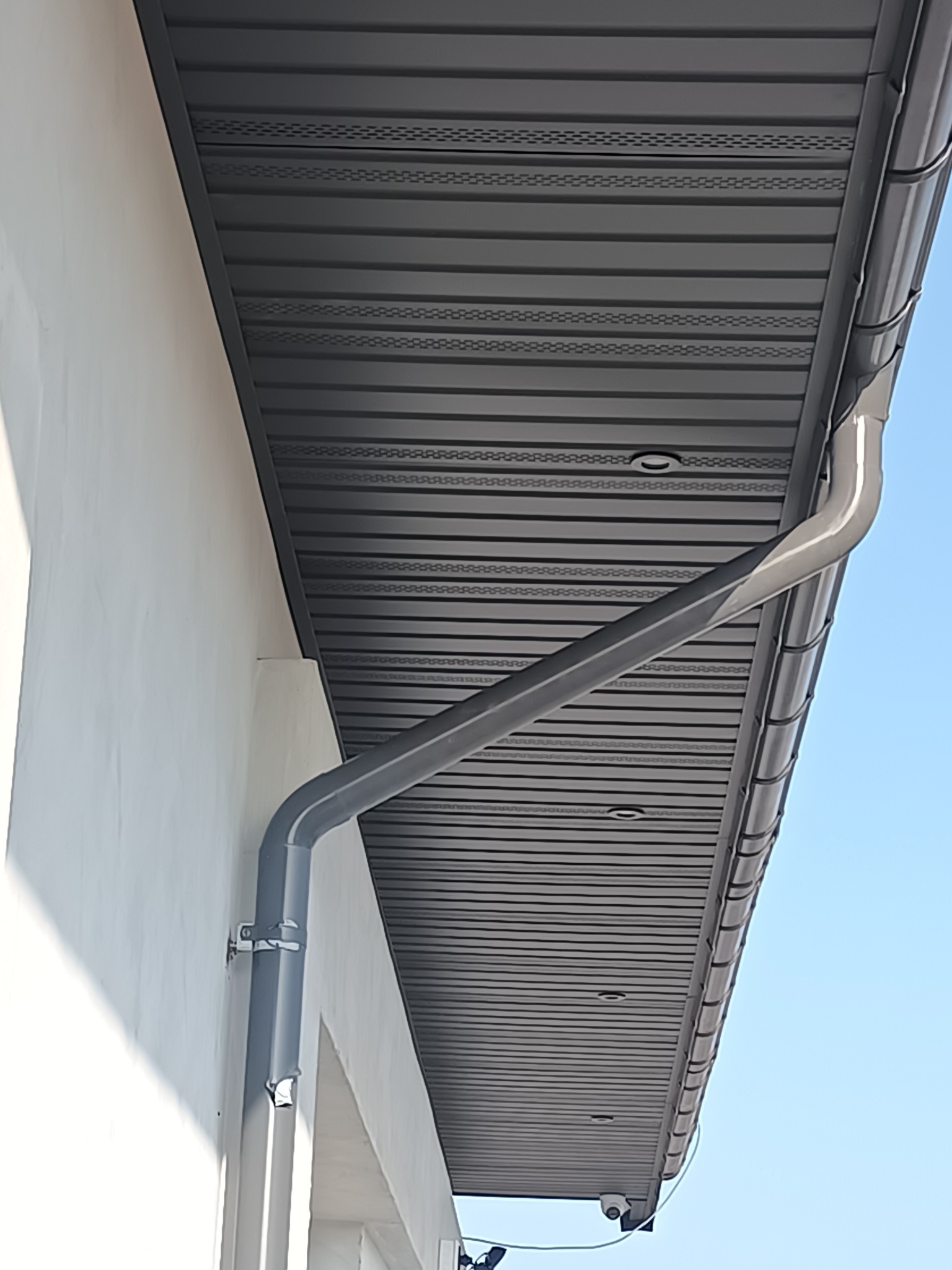 Szara rynna pionowa przymocowana do białej ściany budynku, z widocznym podbitką dachową w tle i fragmentem niebieskiego nieba.