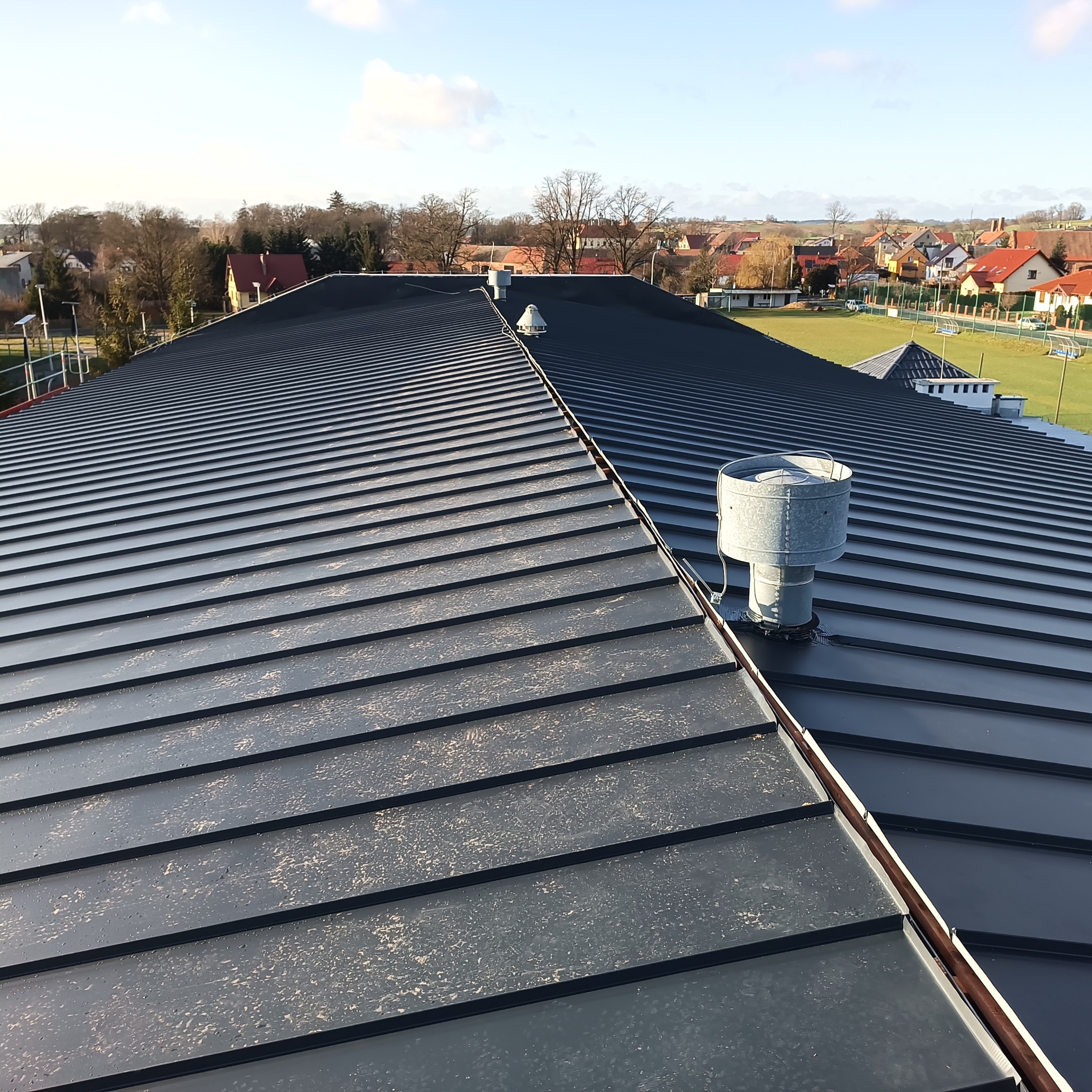 Nowoczesny, grafitowy dach z blachy na budynku z wentylacją kominkową. W tle zabudowa jednorodzinna i błękitne niebo. Widok z góry.