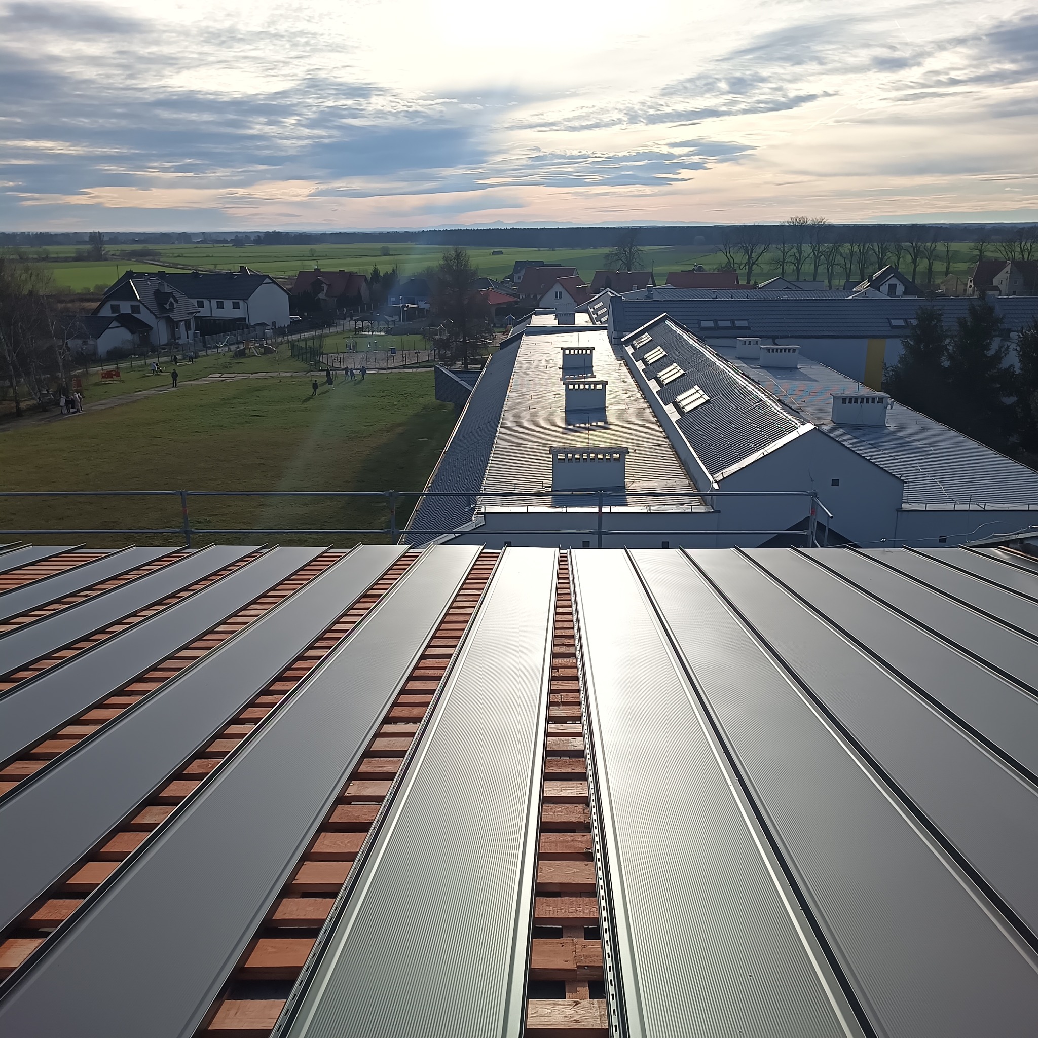 Nowe, szare panele dachowe z widoczną konstrukcją drewnianą w trakcie montażu. W tle budynki mieszkalne i krajobraz wiejski pod błękitnym niebem.