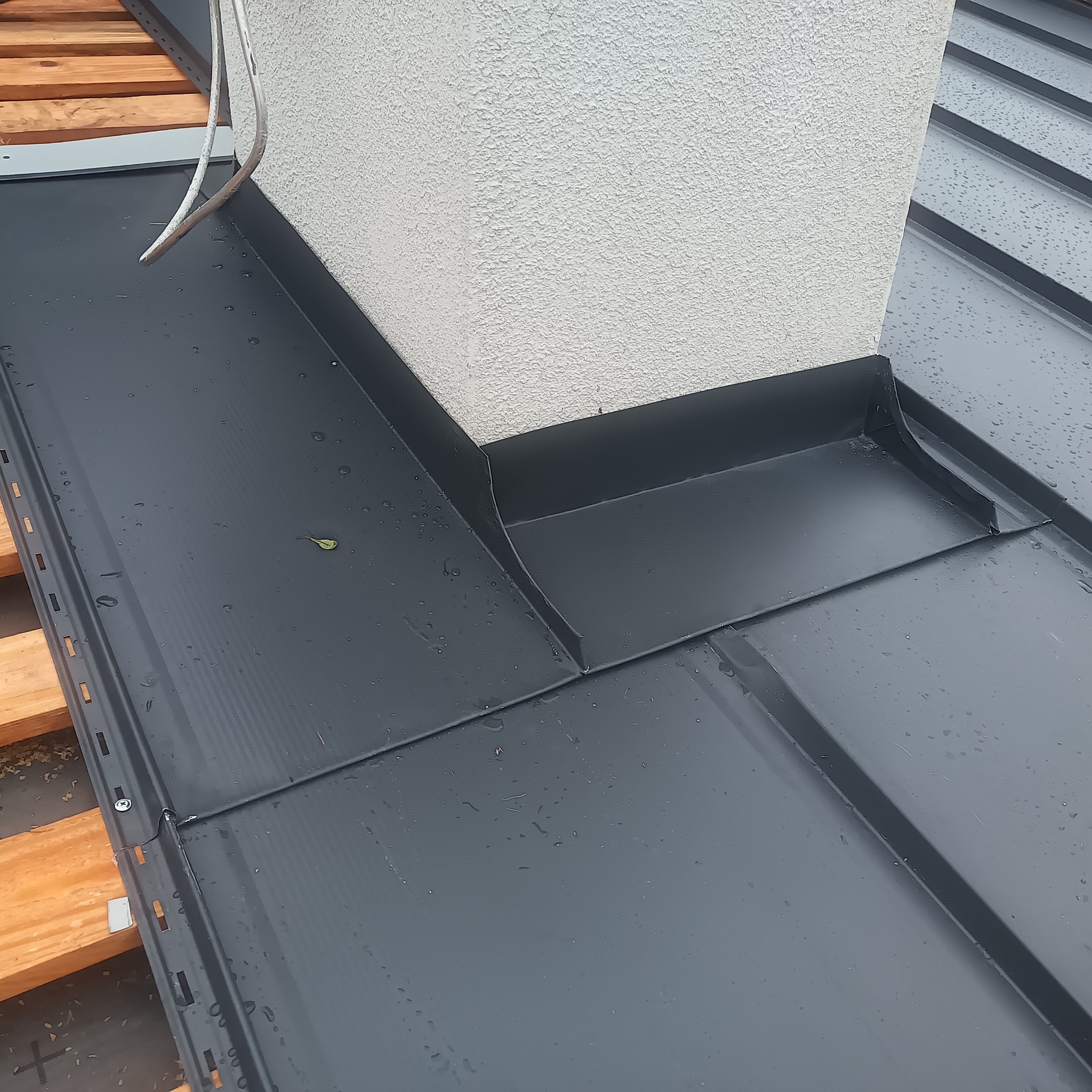 Szczegółowe ujęcie obróbki blacharskiej komina na dachu z blachy na rąbek stojący, widoczne krople wody i fragment drewnianej konstrukcji dachu.