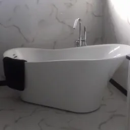 Wolnostojąca, biała wanna z chromowaną armaturą w łazience wykończonej białym marmurem, z czarnym ręcznikiem na krawędzi.