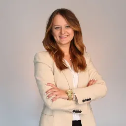 Portret uśmiechniętej kobiety w beżowym żakiecie, białej koszuli i złotym zegarku, stojącej na jasnym tle z założonymi rękami.