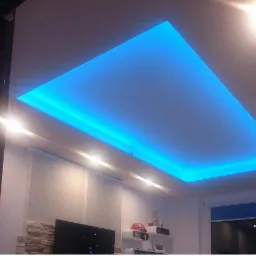 Podwieszany sufit z niebieskim oświetleniem LED w kształcie prostokąta, widok od dołu, fragment ściany z płyt gipsowych i telewizor w tle.