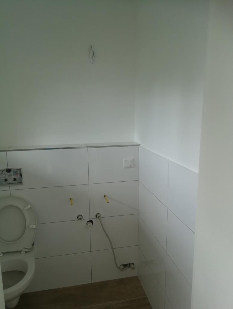 Biała łazienka w trakcie prac wykończeniowych, widoczne płytki na ścianach, otwarta toaleta, wystające rurki i kable, surowe wykończenie.