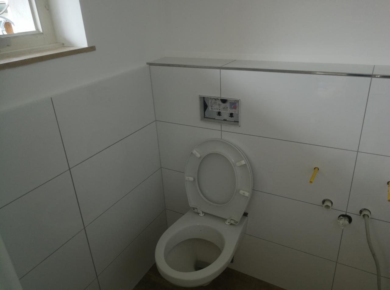 Biała toaleta z otwartą klapą na tle białych płytek, widoczne rury i otwór na przycisk spłukiwania, minimalistyczny wystrój.