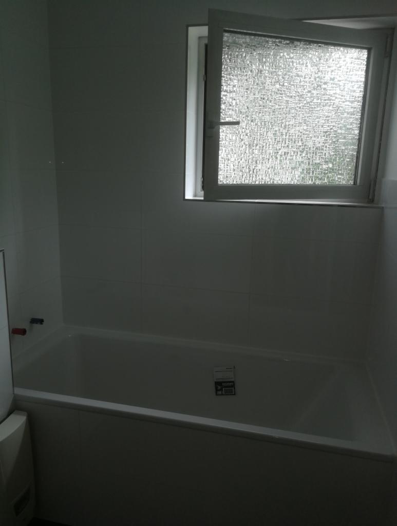 Wanna obudowana białymi płytkami, z otwartym oknem z szybą ornamentową w tle, widoczne baterie i naklejki na wannie.