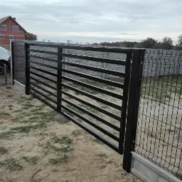Nowoczesna, czarna brama z poziomych profili, przylegająca do ogrodzenia z siatki zgrzewanej na betonowej podmurówce, na tle szarego nieba i częściowo widocznego domu z czerwonej cegły.