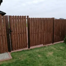 Brązowe ogrodzenie z pionowych desek z furtką, osadzone na ceglanej podmurówce, widok z trawnika w pochmurny dzień.