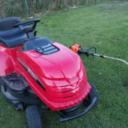 Czerwony traktorek do koszenia trawy z kosiarką żyłkową leżącą obok na trawniku.