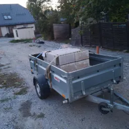 Przyczepka samochodowa załadowana kostką brukową, zaparkowana na żwirowej nawierzchni w pobliżu posesji z nowym chodnikiem.