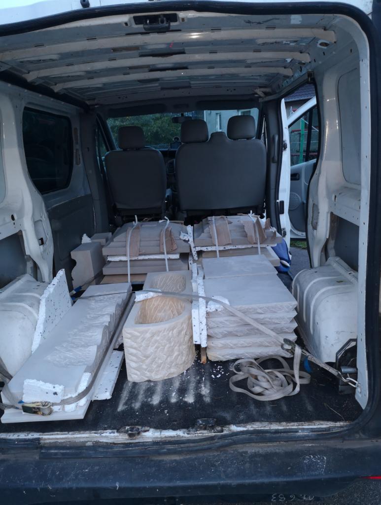 Wnętrze białego busa wypełnione elementami z jasnego kamienia, prawdopodobnie fragmentami fontanny, donicami i obrzeżami zabezpieczonymi pasami transportowymi.