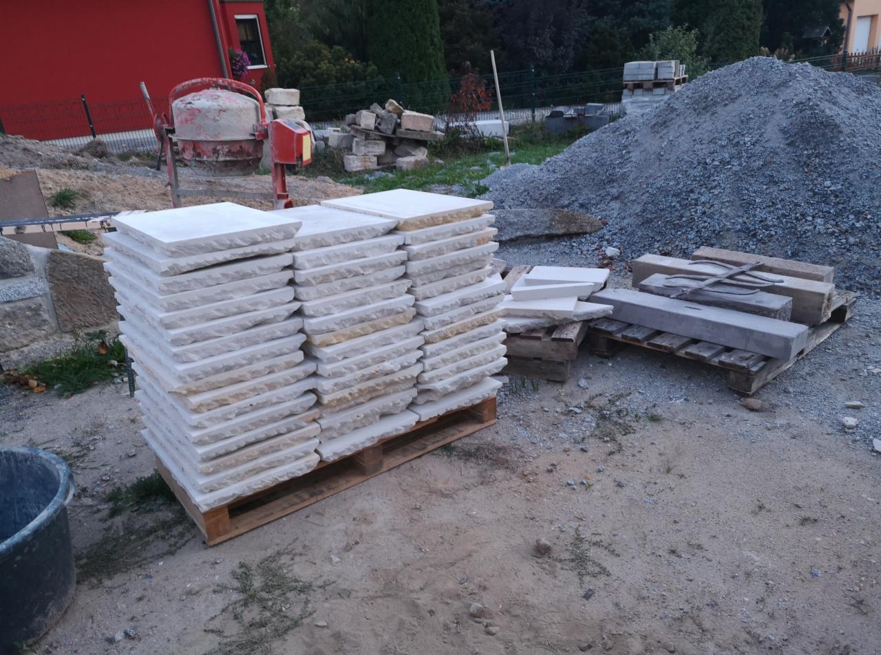 Stos płyt chodnikowych z piaskowca ułożonych na palecie, obok pryzma żwiru i betonowe elementy, w tle czerwony budynek i betoniarka.