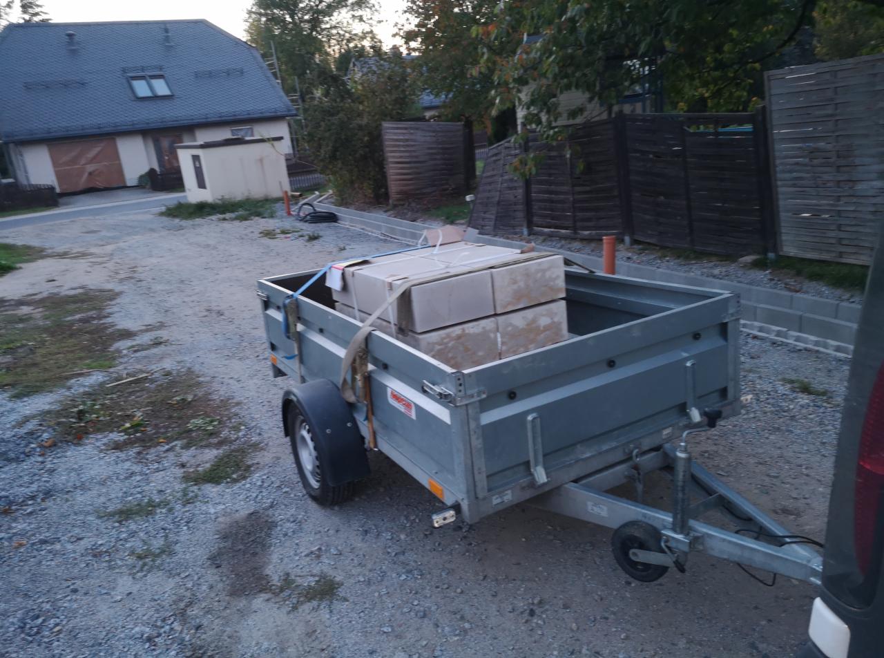 Przyczepka samochodowa załadowana kostką brukową, zaparkowana na żwirowej nawierzchni w pobliżu posesji z nowym chodnikiem.