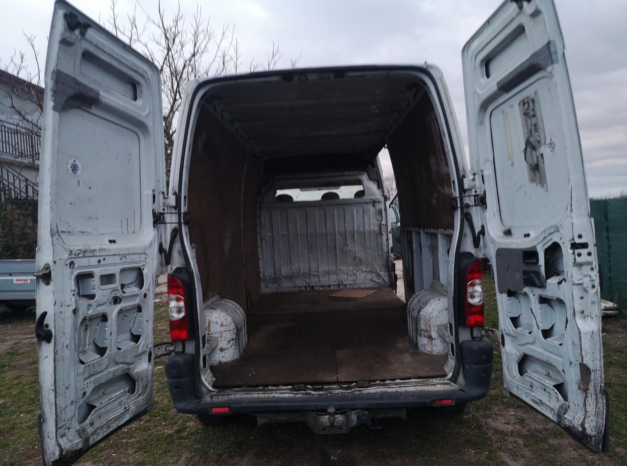 Wnętrze paki dostawczego busa z otwartymi drzwiami, widoczne ślady użytkowania i brudna podłoga, na zewnątrz widoczne drzewa bez liści i fragment zabudowań.