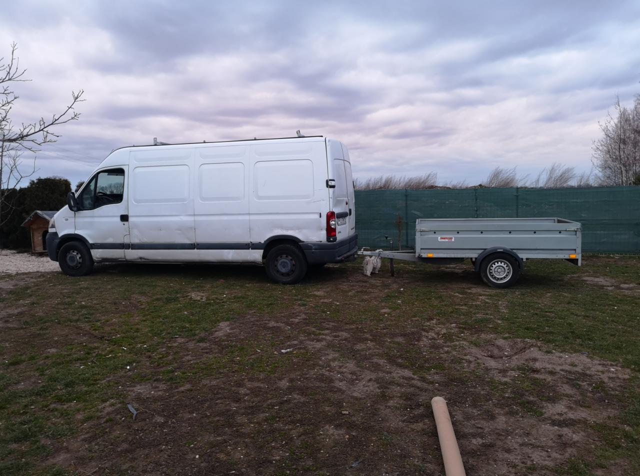 Biały van z przyczepką na trawiastym terenie pod pochmurnym niebem.