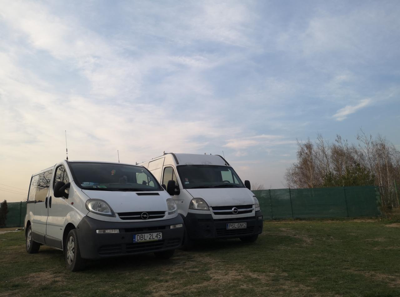 Dwa białe busy Opel Vivaro zaparkowane na trawiastym terenie, widoczne tablice rejestracyjne DBL 2L45 i PSL GN12, tło stanowi błękitne niebo z chmurami i zielone ogrodzenie.
