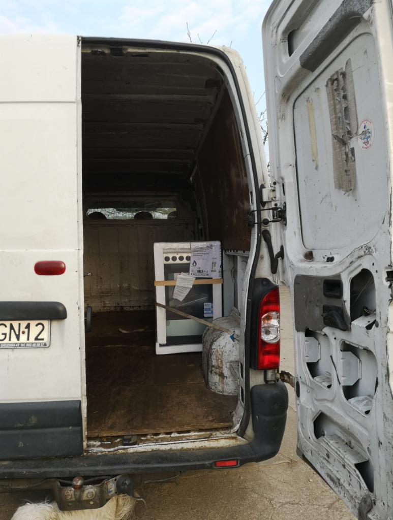 Biały bus z otwartymi tylnymi drzwiami, wewnątrz zapakowana w karton nowa kuchenka, widoczna tablica rejestracyjna pojazdu.