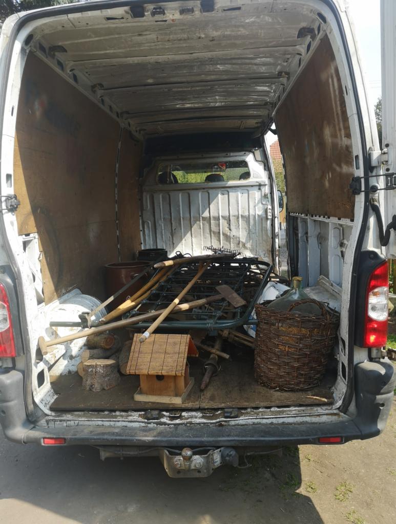 Wnętrze brudnego, białego busa wypełnione różnorodnymi narzędziami ogrodniczymi i akcesoriami, w tym grabiami, łopatami, drewnianym karmnikiem dla ptaków i butlą w wiklinowym koszu.