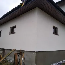 Fragment otynkowanej elewacji domu jednorodzinnego z czterema oknami, widoczne narzędzia budowlane i drewniane podpory.