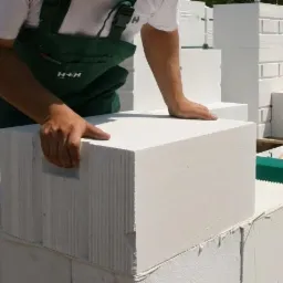 Murarz w zielonym kombinezonie układa biały bloczek z betonu komórkowego na ścianie, widoczne narzędzia murarskie i zapas bloczków.