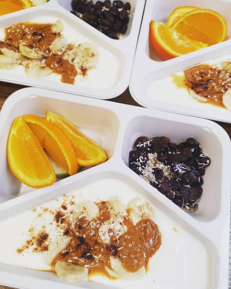 Podzielone białe tacki z jogurtem, bananami, masłem orzechowym, cynamonem, rodzynkami, nasionami słonecznika i pomarańczami, ułożone na drewnianym stole.