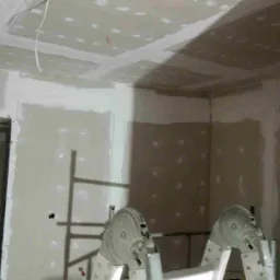 Wnętrze w trakcie prac wykończeniowych: ściany i sufit pokryte płytami gipsowo-kartonowymi, widoczne miejsca łączeń wymagające szpachlowania, drabina aluminiowa w pomieszczeniu, zabezpieczone folią...