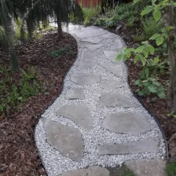 Kręta ścieżka w ogrodzie wyłożona nieregularnymi płytami kamiennymi i drobnym białym żwirem, obrzeżona czarnym profilem, otoczona korą, zielenią i drzewami.