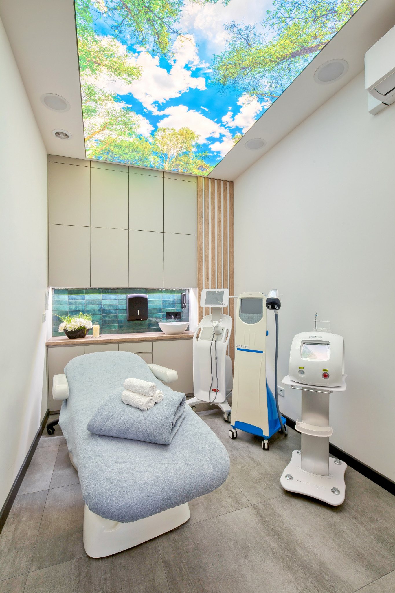 Jasne wnętrze gabinetu z łóżkiem do zabiegów, trzema ręcznikami, umywalką oraz specjalistycznymi urządzeniami kosmetycznymi; sufit z fototapetą imitującą niebo i korony drzew.